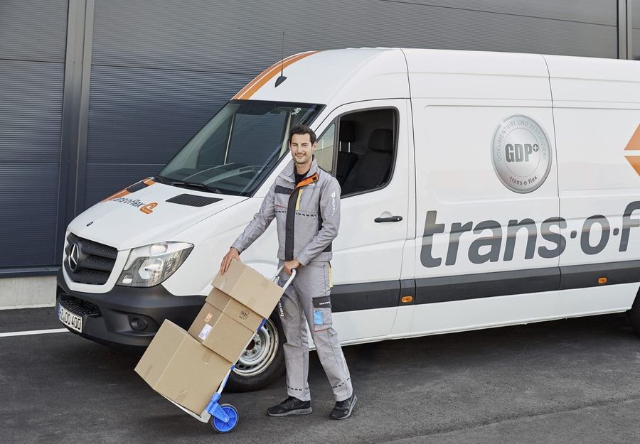 Ein Zustellfahrzeug von dem Logistikdienstleister trans-o-flex. Davor ist ein Paketzusteller mit einer Sackkarre und darauf drei Paketen zu sehen.